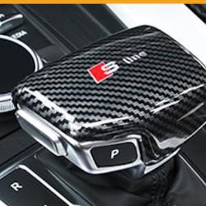 May include: A carbon fibre gear shift knob with a silver base and a red "S line" logo. The knob has a textured, woven pattern and a button labelled "P". The background shows part of a car's console.