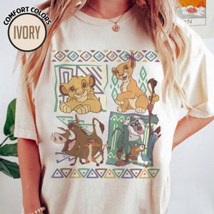 May include: Ivory-colored t-shirt featuring a colorful graphic of characters from The Lion King. The design includes Simba, Nala, Timon, Pumbaa, Rafiki, and Zazu, set against geometric patterns in green, blue, and yellow. The shirt has the words "Comfort Colors" and "Ivory" on it.