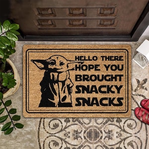 May include: Rectangular doormat with a black and tan design. The mat features a black silhouette of a character with the text "HELLO THERE HOPE YOU BROUGHT SNACKY SNACKS" in black. The mat is placed in front of a brown door.