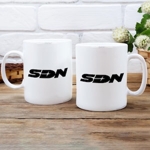 May include: Two white ceramic mugs with the black letters "SDN" printed on them. The mugs are on a wooden surface with a white brick background. The mug on the left has the word "FRONT" printed on it. The mug on the right has the word "BACK" printed on it.