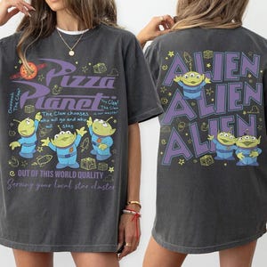 May include: A charcoal gray t-shirt featuring a Pizza Planet graphic on the front with three green aliens, planets, and text including "The Claw chooses who will go and who will stay." The back displays "ALIEN" in large purple letters with aliens.