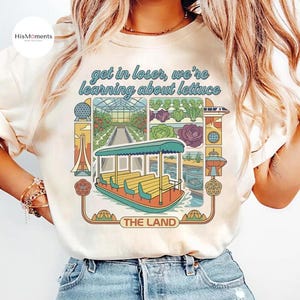 May include: Cream-colored t-shirt featuring a colorful graphic with the text "get in loser, we're learning about lettuce." The graphic includes a boat, greenhouse, and various vegetables. The words "THE LAND" are also visible.