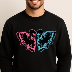 Op de afbeelding: Zwart sweatshirt met een afbeelding van twee gamers met zonnebrillen, één in roze en de andere in blauw, die gamecontrollers vasthouden. Het ontwerp is een gestileerde illustratie, voor gamers.