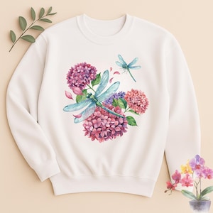 May include: A cream-colored crewneck sweatshirt featuring a central watercolor-style graphic of two blue-winged dragonflies hovering over clusters of pink and purple hydrangea flowers, with scattered pink petals surrounding the floral arrangement.