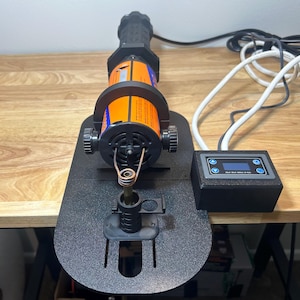May include: An industrial tool, black and orange in colour, with a handle, cylindrical body, and base. It features a heating element and is connected to a control module with a digital display. The tool is on a wooden surface.