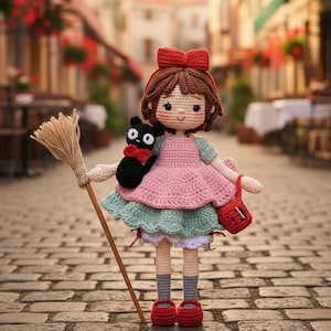 Puede incluir: Muñeca de ganchillo con un vestido rosa con falda verde, zapatos rojos y un lazo rojo. La muñeca sostiene una escoba y un gato negro con pajarita roja. Un bolso rojo cuelga del brazo de la muñeca.