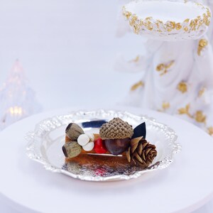 May include: A silver-colored decorative plate holds a miniature arrangement of natural elements. The arrangement includes an acorn, pinecone, and other decorative items. A white angel figurine and two illuminated Christmas tree decorations are in the background.