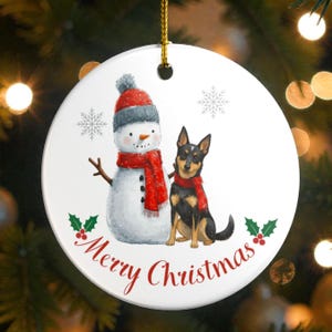 May include: A round white ceramic Christmas ornament featuring a printed illustration of a snowman wearing a red hat and scarf standing next to a black and tan dog wearing a red scarf. The text Merry Christmas is printed in red script below.