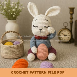 Op de afbeelding: Een gehaakte konijnenpop met gesloten ogen, gekleed in een blauwe trui en een bordeauxrode broek, met een roze ei in de hand. Een mand met kleurrijke eieren en een oranje wortel staan in de buurt. De tekst "CROCHET PATTERN FILE PDF" staat onderaan.