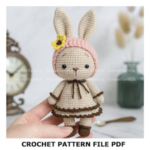Pode incluir: Uma boneca de coelho de crochê usando um chapéu rosa com uma flor amarela, um vestido bege com acabamento marrom e sapatos marrons. A boneca tem orelhas longas e um laço marrom. O texto "CROCHET PATTERN FILE PDF" está na parte inferior.