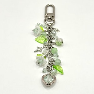 May include: A silver-toned keychain with a lobster clasp, adorned with green and clear beads, leaf charms, star charm, and flower charms. A heart-shaped charm hangs at the bottom. The charms are connected by a silver chain.