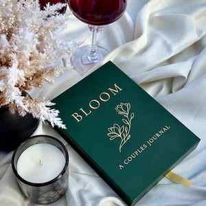 May include: A dark green journal with gold lettering that reads "BLOOM" and "A COUPLES JOURNAL" with a floral illustration. A lit candle in a glass holder, two wine glasses filled with red wine, and decorative white flowers are also present.