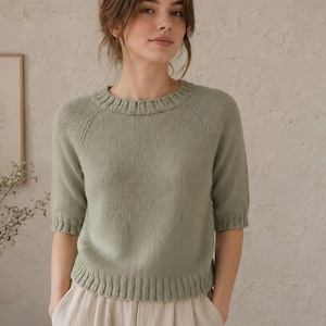 May include: A sage green knit jumper featuring a round neckline, raglan sleeves, and ribbed detailing at the collar, cuffs, and hem. The jumper has a smooth, stockinette stitch texture and reaches to the waist, paired with light beige trousers.