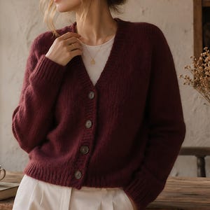 May include: A deep burgundy V-neck cardigan featuring a button-down front with four dark buttons, long sleeves with ribbed cuffs, and a ribbed hem. The cardigan is worn over a plain white shirt, creating a layered look with a textured knit finish.