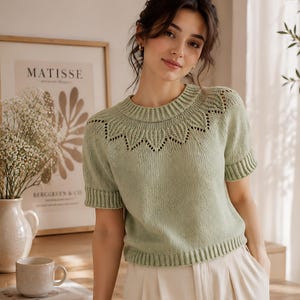 May include: A light sage green short-sleeved knit sweater featuring a round ribbed neckline and a decorative eyelet pattern around the yoke. The garment has ribbed cuffs and a ribbed hem, crafted with a smooth stockinette stitch texture throughout.