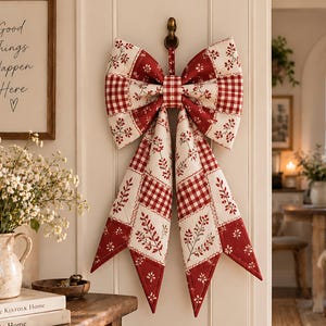 May include: A large, decorative fabric bow hanging on a wall hook. The bow is constructed from quilted panels featuring a red and white gingham check pattern alternating with a cream-colored fabric printed with small red floral sprigs and leaves.