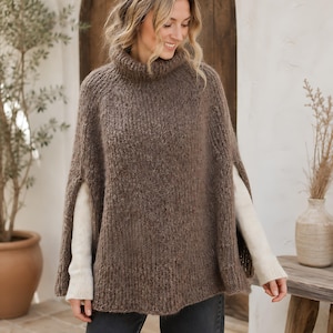 May include: A brown knit poncho featuring a high turtleneck collar and open sides, worn over a cream-coloured long-sleeve sweater and dark grey jeans. The garment has a textured, fuzzy appearance with ribbed detailing along the collar and hemline.