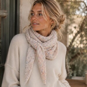 May include: A cream-colored long-sleeve sweater paired with a triangular knit scarf. The scarf features a speckled, multi-colored yarn pattern with flecks of blue, orange, and pink throughout the light beige base, tied in a simple front knot.