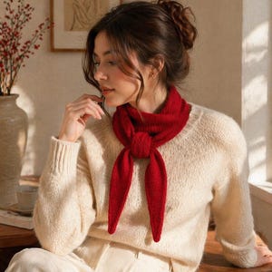 Puede incluir: Una mujer con un suéter color crema y pantalones blancos, con una bufanda roja brillante atada al cuello. Los extremos de la bufanda cuelgan. Un jarrón con bayas rojas y una impresión enmarcada están en el fondo.