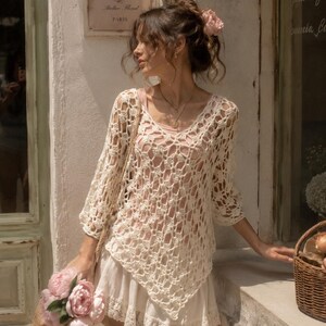 May include: Cream-colored, open-weave crochet top with a V-neck, paired with a white skirt. The image shows a woman holding a bag of pink flowers. A sign that says "LA VIE EN ROSE Atelier Floral PARIS" is visible.