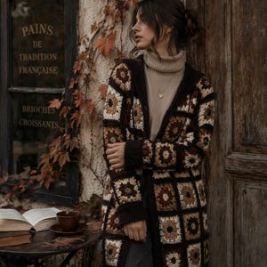 Può includere: Una donna indossa un cardigan all'uncinetto marrone, crema e nero con un dolcevita. L'immagine mostra una scena di caffè con un tavolo, libri e una tazza di caffè. La finestra recita "PAINS DE TRADITION FRANCAISE" e "BRIOCHES CROISSANTS".