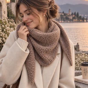 May include: A textured, triangular knit scarf in a muted brown shade, worn over a cream-coloured coat. The scarf features a dense, bumpy stitch pattern and pointed ends that drape over the shoulders, providing a warm layer over the light coat.