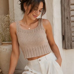 May include: A beige sleeveless knit crop top featuring a square neckline and a wide ribbed hem. The garment is constructed with a stockinette stitch body and a decorative ribbed border along the neckline and armholes, ending at the waistline.