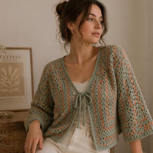 Puede incluir: Cárdigan de crochet verde claro y melocotón con cierre de lazo. El diseño de tejido abierto presenta un cuello en V y mangas tres cuartos. El cárdigan se lleva sobre una camiseta blanca. Una mesita de noche de madera y un estampado enmarcado están en el fondo.