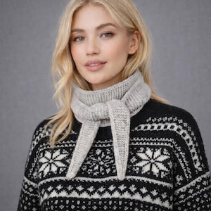 Könnte beinhalten: Hellgrauer Strickschal, der zu einem Knoten gebunden ist und über einem schwarz-weiß gemusterten Pullover getragen wird. Der Pullover hat ein Schneeflocken- und geometrisches Design. Der Schal ist aus weichem, strukturiertem Material.