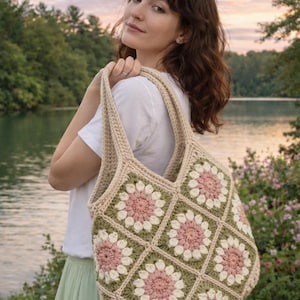 May include: A beige crochet tote bag with a floral pattern in pink, white, and green. The bag has a square design and is carried by a person. The background features a lake and trees.