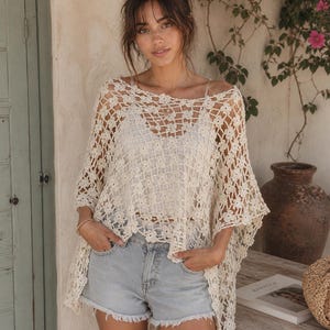 May include: A cream-coloured, open-weave crochet top with a floral pattern. The top is worn over a white vest and paired with light blue denim shorts. The image is taken outdoors with a neutral background.