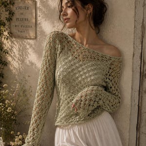 May include: A light green, open-weave long-sleeve top with an off-the-shoulder neckline. The top is paired with a white skirt. A sign that reads "ATELIER DE FLEURS 7, RUE D'ALESIA PARIS" is visible in the background.
