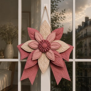 May include: A decorative, quilted flower in shades of pink and beige, hanging from a window. The flower has layered petals and a central, ruffled detail. The design includes a bow at the base, and is suspended by a natural fiber cord.