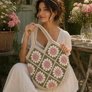 May include: A crochet tote bag constructed from square motifs featuring a floral pattern. The squares alternate between sage green and cream borders, each containing a central pink flower with white petals. The bag has two integrated handles.