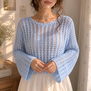 Puede incluir: Una blusa de punto calado azul claro con mangas largas. La blusa tiene un cuello redondo y un corte holgado y aireado. Las mangas son acampanadas en la muñeca. La blusa se lleva sobre una prenda blanca.