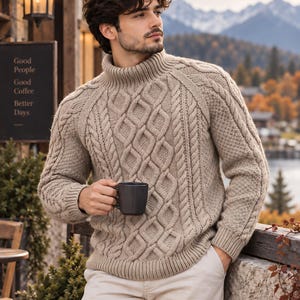 Peut inclure: Un pull à col roulé beige présentant un motif complexe en tricot torsadé sur le devant et les manches, associé à un pantalon clair. Le pull possède un col, des poignets et un ourlet côtelés, et est porté en tenant une tasse gris foncé.