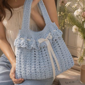 May include: A light blue crochet tote bag featuring a textured shell stitch pattern, two sturdy handles, and a ruffled top edge adorned with a white satin ribbon tied into a bow at the center. The bag is constructed from yarn in a soft pastel hue.