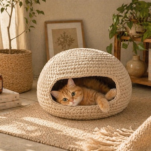 以下が含まれることがあります： ベージュのかぎ針編みの猫用ベッド。丸い形で、入り口があります。オレンジ色のトラ猫が中にいます。ベッドは織りのラグの上にあり、近くにキャンドルと本があります。