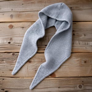 Può includere: Una combinazione di cappuccio e sciarpa in maglia blu chiaro. Il cappuccio è attaccato a una lunga sciarpa con estremità a punta. L'articolo è realizzato in un tessuto a maglia morbido e strutturato. La sciarpa è su una superficie di legno.