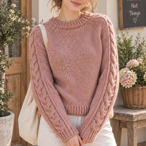 Puede incluir: Un suéter de punto de manga larga en color rosa empolvado con cuello redondo acanalado, puños acanalados y dobladillo acanalado. Las mangas presentan un patrón de trenzas vertical a lo largo de la parte exterior de cada brazo, con pantalones blancos.