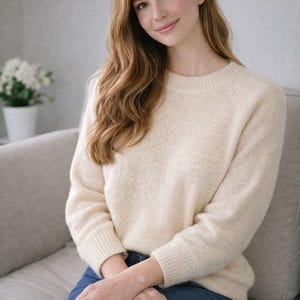 Könnte beinhalten: Cremefarbener Pullover mit Rundhalsausschnitt und langen Ärmeln. Der Pullover wird mit einer blauen Jeans kombiniert. Die Person sitzt auf einem grauen Sofa. Ein weißer Blumentopf ist im Hintergrund.