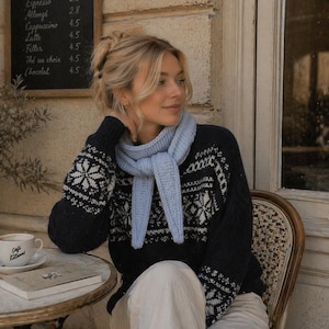 Puede incluir: Una mujer con un suéter azul marino con patrones de copos de nieve blancos y una bufanda azul claro. Está sentada en una mesa de café con una taza de café y un libro. El menú de "Café Kitsuné" está a la vista.