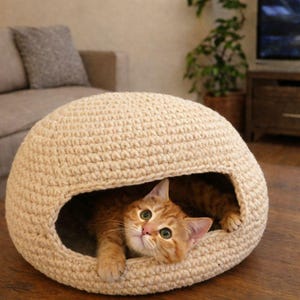 May include: A beige, crocheted cat bed in a dome shape. An orange tabby cat with green eyes is visible in the opening. The cat bed sits on a wooden table, with a sofa and plant in the background.