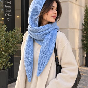 Può includere: Una sciarpa lavorata a maglia color azzurro con un motivo testurizzato a legaccio. La sciarpa ha una forma triangolare che si drappeggia sulla testa e intorno al collo, con due estremità lunghe e appuntite che scendono su un maglione color crema.