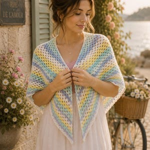 May include: A pastel-colored crochet shawl with blue, yellow, and white stripes. The shawl is worn over a white dress. The background includes a stone wall, flowers, and a bicycle with a basket of flowers.