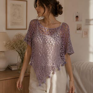 Könnte beinhalten: Ein helllila Häkeltop mit einem markanten Lochmuster und kurzen Ärmeln. Das Top hat einen asymmetrischen Saum und ist mit kleinen Pompons verziert. Es wird über einem weißen Camisole und mit beigefarbenen Hosen getragen.