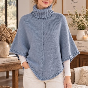 May include: A dusty blue knit poncho featuring a ribbed turtleneck collar, a central vertical seam, and ribbed trim along the hem and sleeve openings. The garment is worn over a white long-sleeved shirt and light-colored trousers.