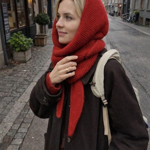 Puede incluir: Una persona con un conjunto de capucha y bufanda de punto rojo, combinado con un abrigo marrón oscuro. La capucha enmarca la cara y la bufanda cae por delante. El conjunto es adecuado para el clima frío. El fondo muestra una calle europea.