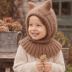Könnte beinhalten: Eine hellbraune Stricksturmhaube mit zwei kleinen spitzen Ohren an der Oberseite und einem gerippten Kragen, der über die Schultern reicht. Die Kopfbedeckung ist aus weichem, strukturiertem Garn gefertigt und bedeckt Kopf und Hals vollständig.