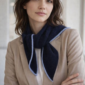 Può includere: Una sciarpa blu navy con un bordo azzurro è drappeggiata intorno al collo. È indossata sopra una camicia bianca e un blazer beige. La donna ha i capelli scuri e guarda la fotocamera.